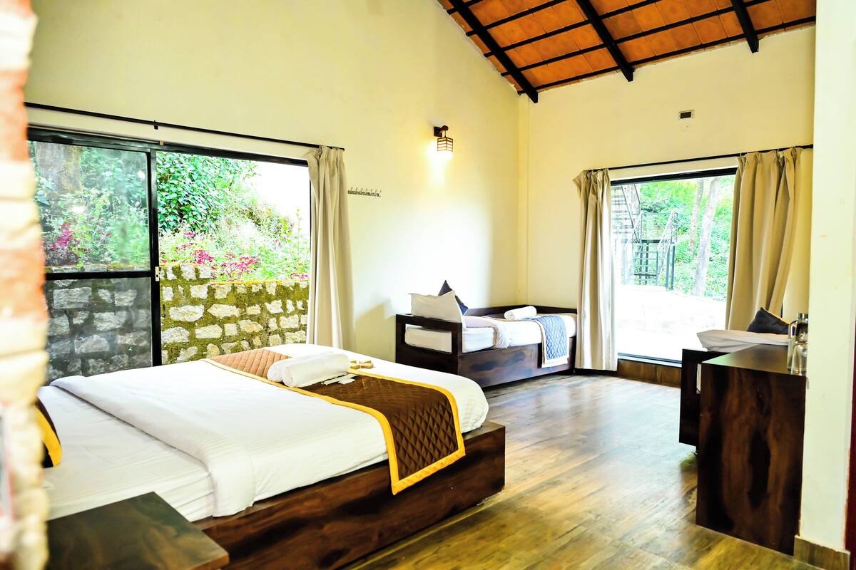 A bedroom with a large bed, a desk, and a view of greenery through the windows.