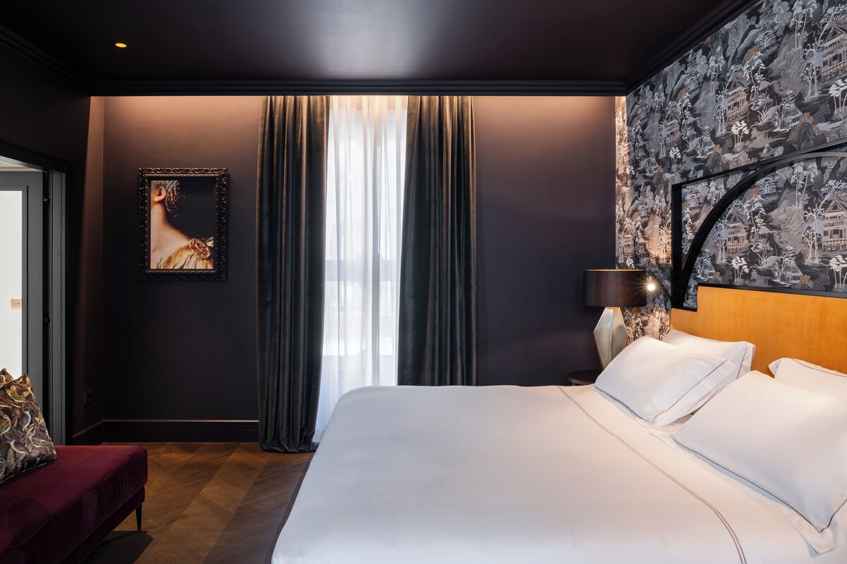 A modern hotel room with a large bed, a red bench, a framed artwork, and a patterned wallpaper.