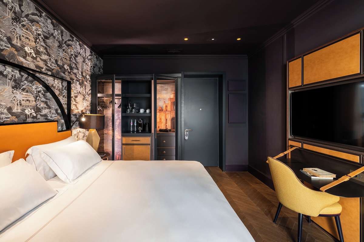 A hotel room with a large bed, a desk with a chair, a TV, and a wardrobe.