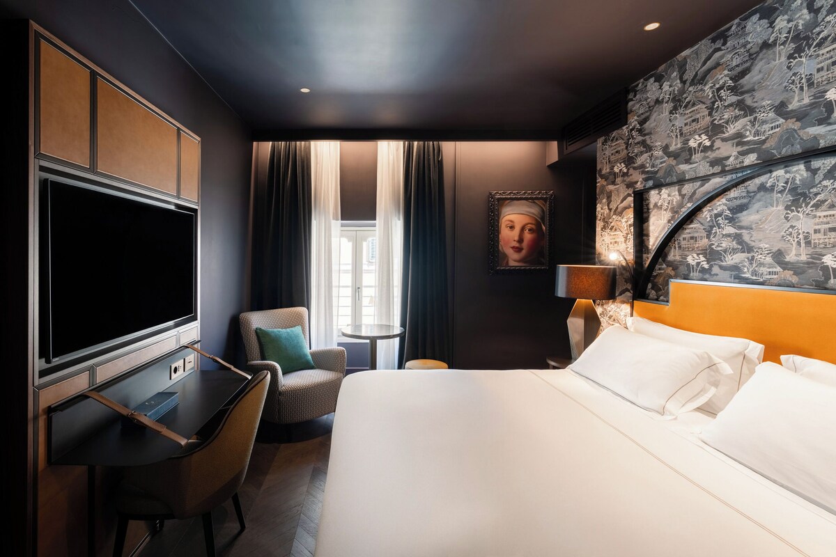 A hotel room with a bed, a desk, a chair, a TV, and a small table.