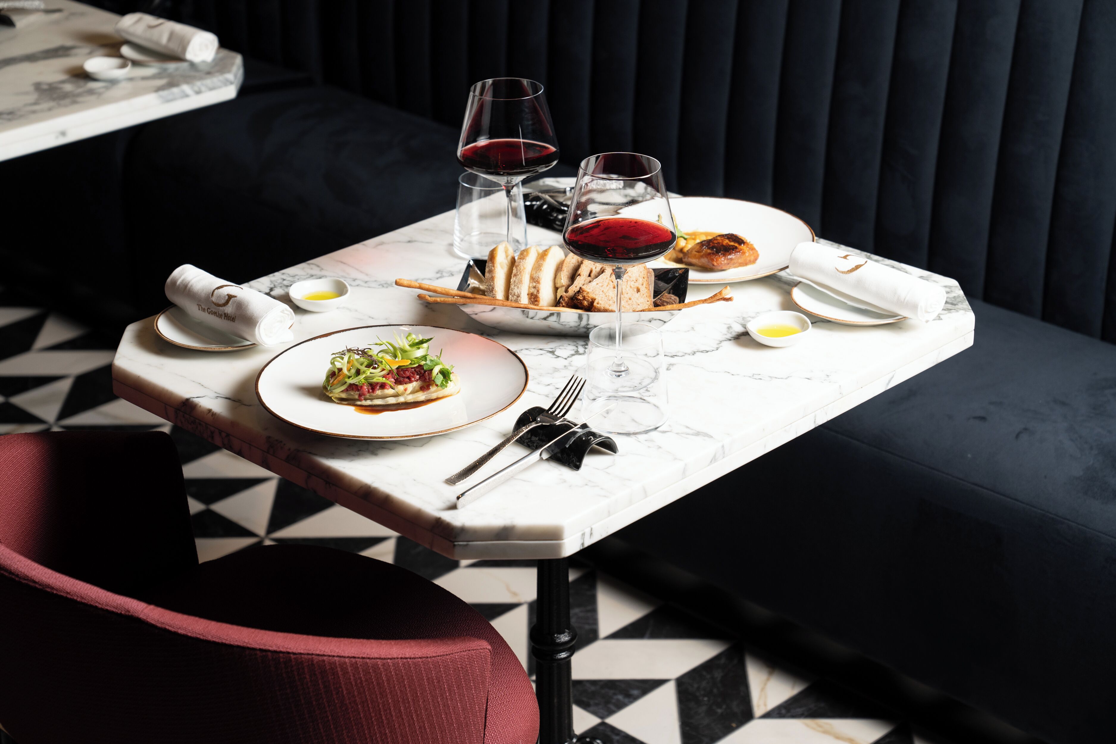 A dining table with plates of food, glasses of red wine, and a napkin with a logo.