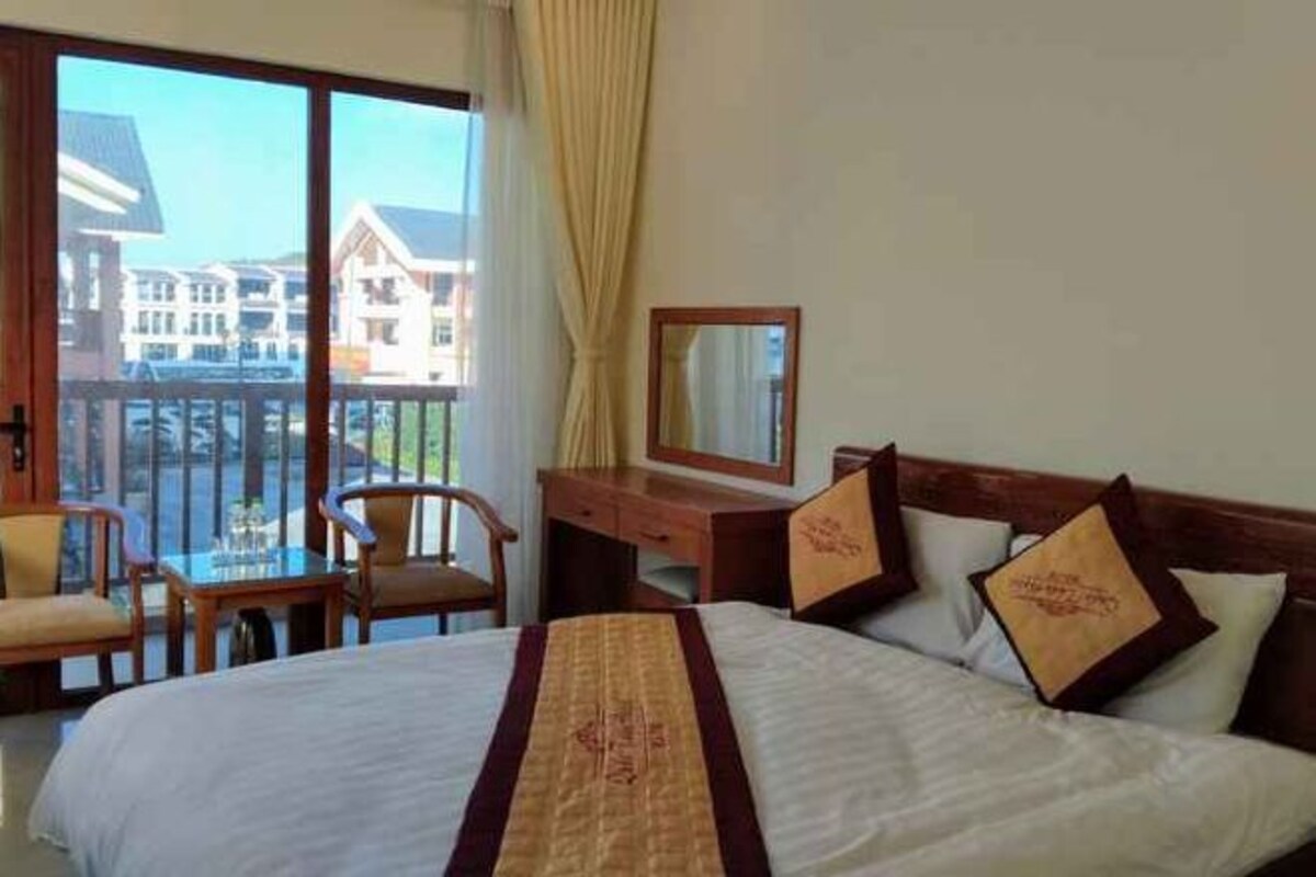 A hotel room with a bed, a desk, a chair, and a balcony with a view of buildings.