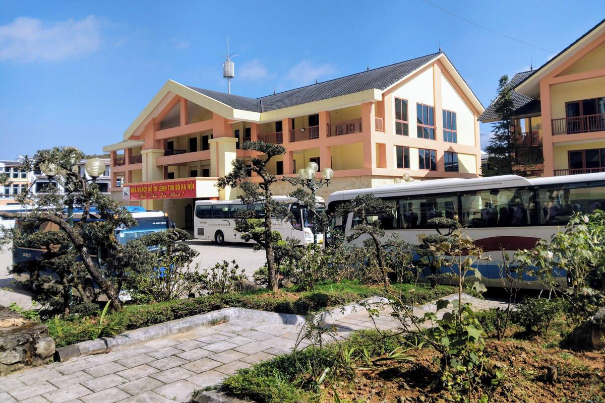 Un immeuble à plusieurs étages, doté d’un toit plat et de balcons. Un autobus est garé devant, et une enseigne avec du texte dans une langue étrangère est visible.
