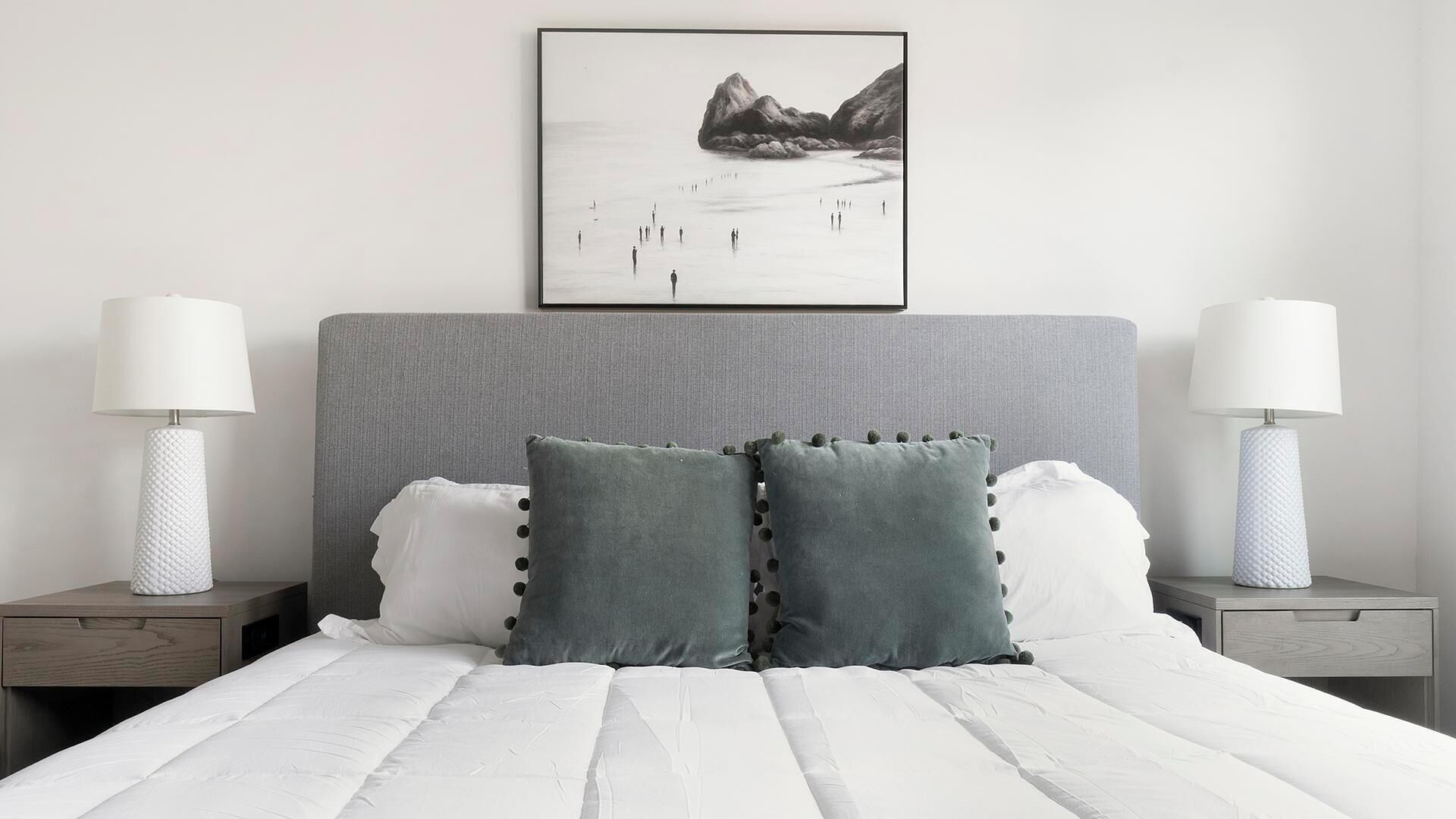 A neatly made bed with white linens, two grey pillows, and a grey headboard. There are two bedside tables with lamps and a framed picture above the bed.