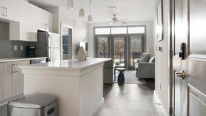 A modern kitchen with white cabinetry, a built-in island, and a dining area with a gray sofa and a small table. - Landing | Spectacular 2BD, Pool, Gym (Atlanta)