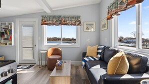 A modern living room with a sofa, armchair, and a view of a lake through large windows. - Cozy Waterfront Cottage (Noank)