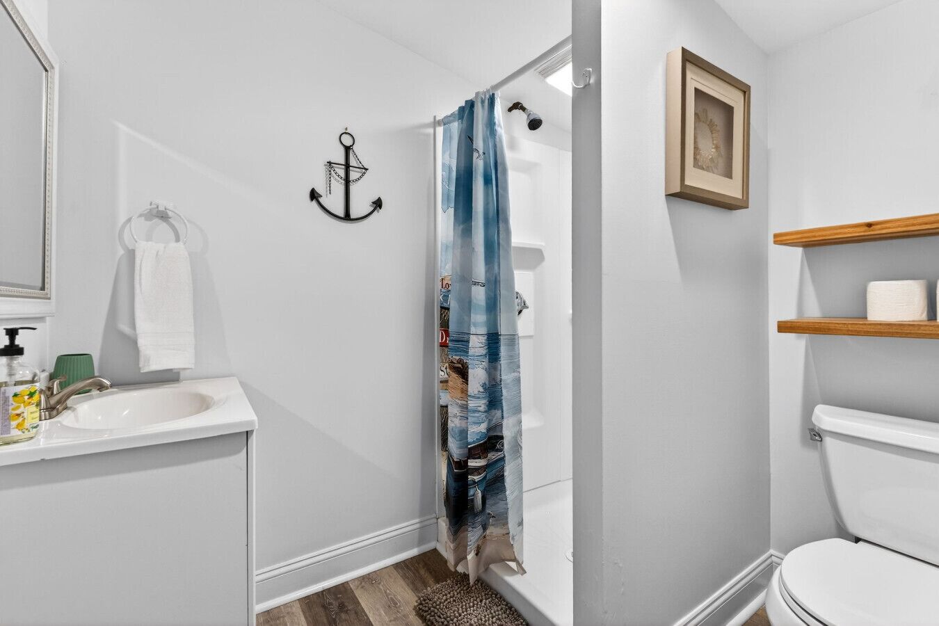 Ein Badezimmer mit einem weißen Waschbecken, einer Duschvorhang, einem Holzregal mit Toilettenpapier und einem gerahmten Bild an der Wand.