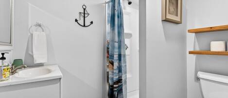 Ein Badezimmer mit einem weißen Waschbecken, einer Duschvorhang, einem Holzregal mit Toilettenpapier und einem gerahmten Bild an der Wand.