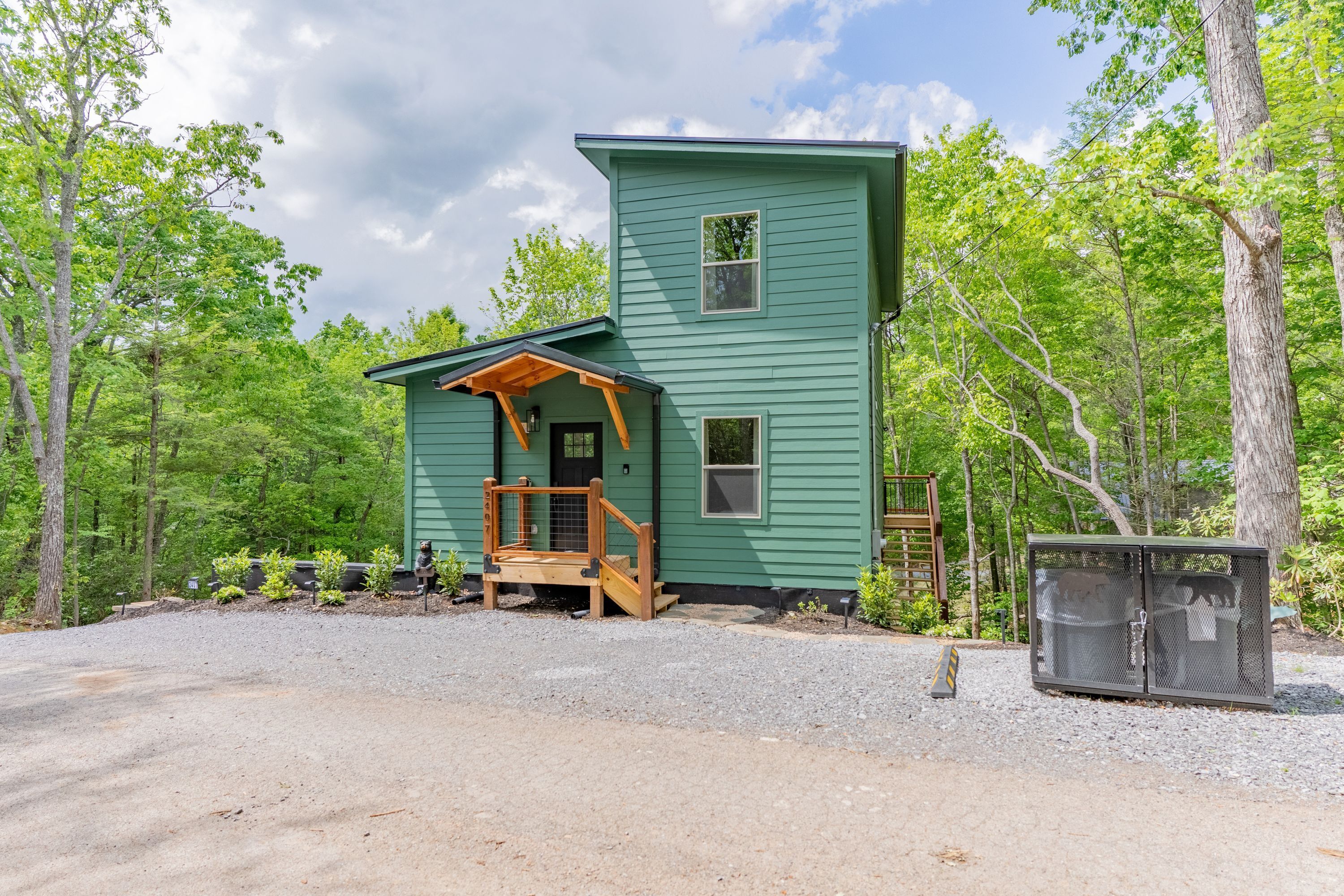 A two-story cabin with a porch and a fenced-in area, surrounded by trees.
