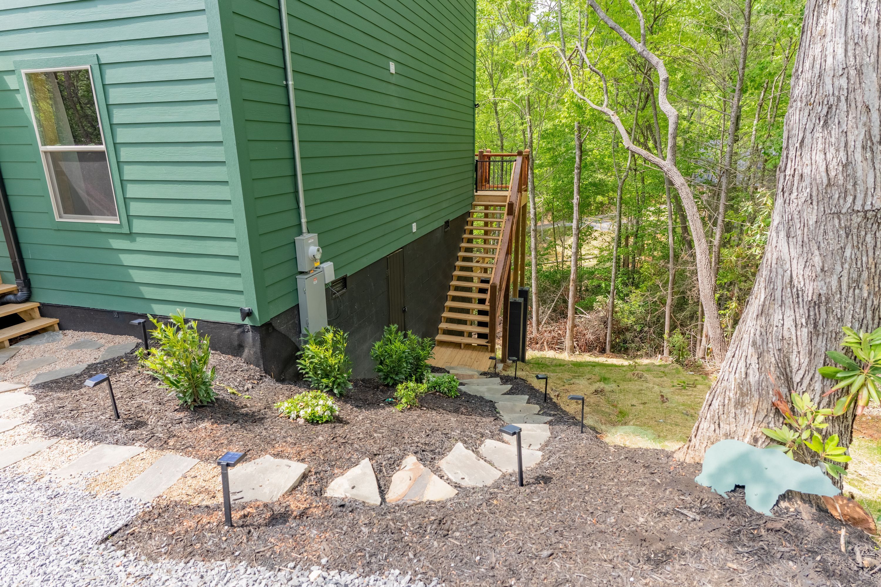 Una casa verde con una escalera de madera que lleva a una terraza, rodeada de un jardín con piedras para caminar y plantas pequeñas.