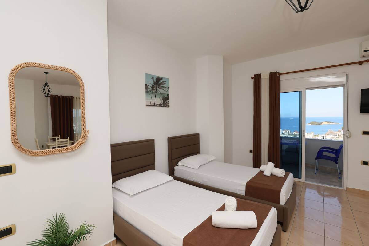 A hotel room with two beds, a large mirror, a potted plant, a painting, and a view of the sea through a sliding door.