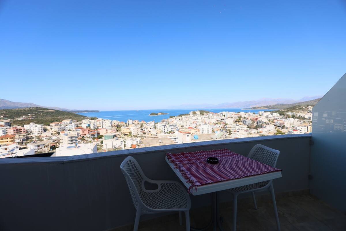A balcony with a table and two chairs, overlooking a coastal town with buildings and mountains in the distance.
