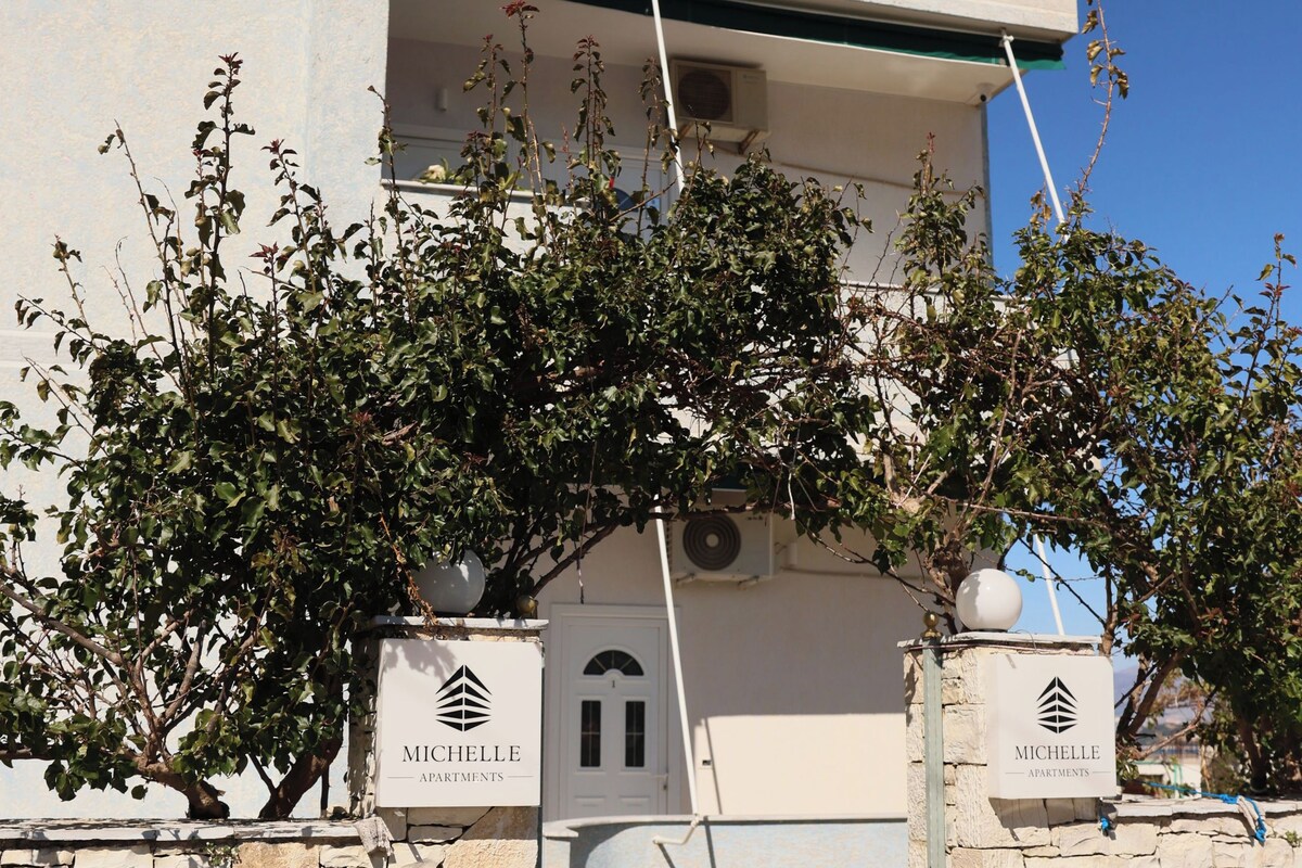 A modern building with a stone facade and a balcony. There are two signs with the name "MICHELLE APARTMENTS".