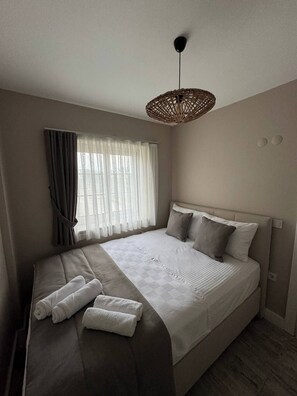 Una cama bien hecha con almohadas y una colcha, una mesita de noche con toallas, una ventana con cortinas y una lámpara colgante.