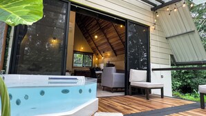 Un moderno espacio exterior con jacuzzi, un muelle de madera, una zona de estar con una silla y una mesa, y un amplio ventanal que ofrece vistas del entorno verde.