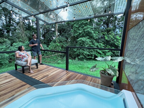 Een houten terras met een hottub, omringd door groen en een metalen pergola met lichtslingers.