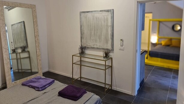 A bedroom with a bed, a bedside table, a mirror, a painting, and a door leading to another room. - Be Magenta Art Home - Yellow style (Magenta)