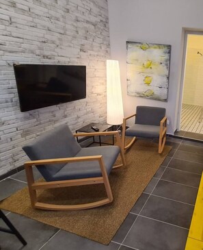 Une chambre moderne avec un mur en briques, un téléviseur à écran plat, deux fauteuils à bascule, une table d’appoint et un grand tableau abstrait.