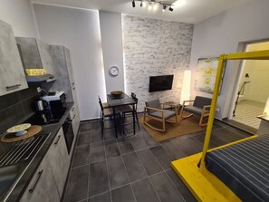 A compact living space with a kitchenette, a dining area, a TV mounted on a brick wall, and a bed with a yellow frame.