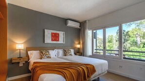 Una cama doble con una manta marrón, dos mesitas de noche con lámparas, un cuadro en la pared y un ventanal con vistas a árboles.