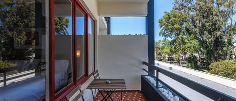 Un balcón con una mesa y banco de madera, una ventana con marco rojo y vista a árboles y una carretera.