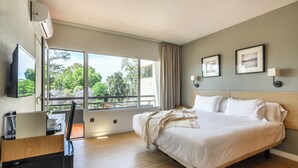 Habitación de hotel con cama, escritorio con una computadora portátil, televisor y balcón con vista a follaje.