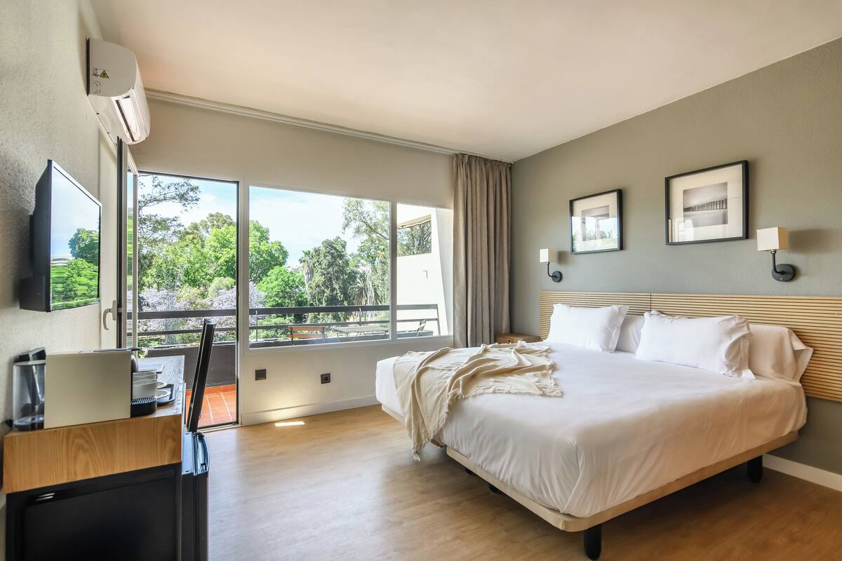 A hotel room with a bed, a desk with a laptop, a TV, and a balcony with a view of greenery.