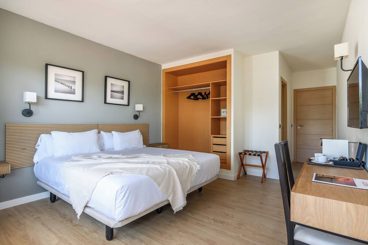 A neatly arranged bedroom with a large bed, a wooden wardrobe, a desk with a chair, and framed pictures on the wall.