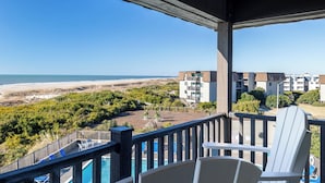 A balcony with a view of the beach and ocean, a pool, and a chair.