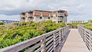 A wooden boardwalk leading to a multi-story building with balconies, surrounded by greenery.