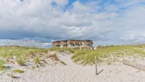 A beachfront building with a sandy path leading to it, surrounded by grass and dunes.