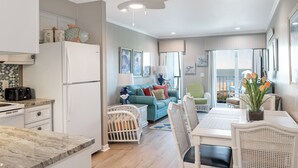A modern kitchen with white appliances, a dining area with white chairs, and a living room with a teal sofa.