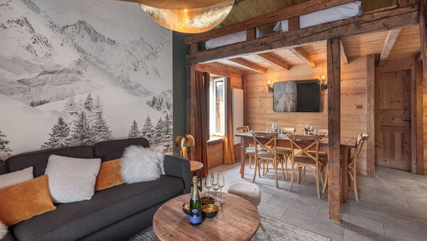 Une pièce chaleureuse avec un plafond en bois, une fresque représentant des montagnes, un canapé avec des coussins, une table à manger avec des chaises, et une petite table ronde sur laquelle se trouvent une bouteille et des verres.