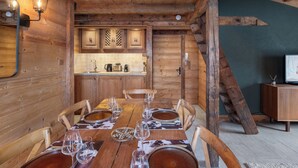 Un intérieur en bois comprenant une table à manger pour six personnes, des chaises en bois, un coin cuisine avec des placards et un téléviseur posé sur un meuble en bois.