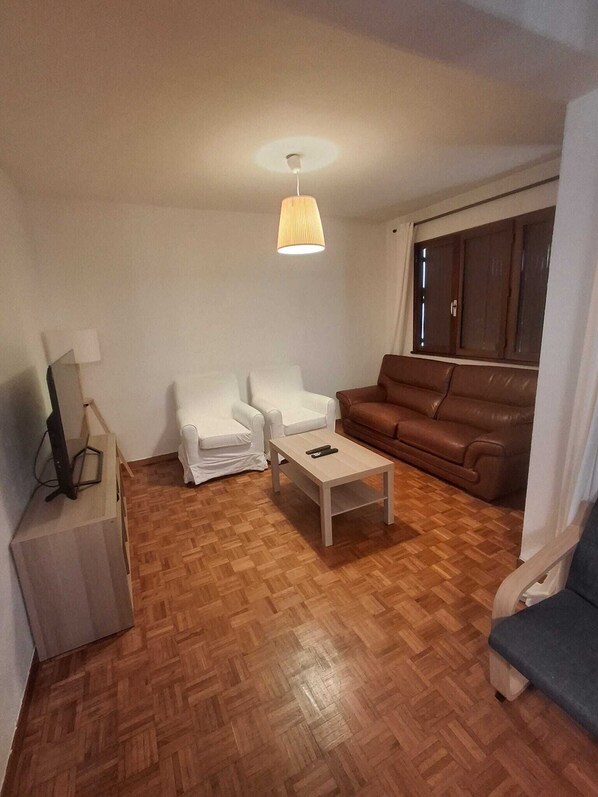 A living room with a brown leather sofa, two white armchairs, a wooden coffee table, and a TV on a stand.