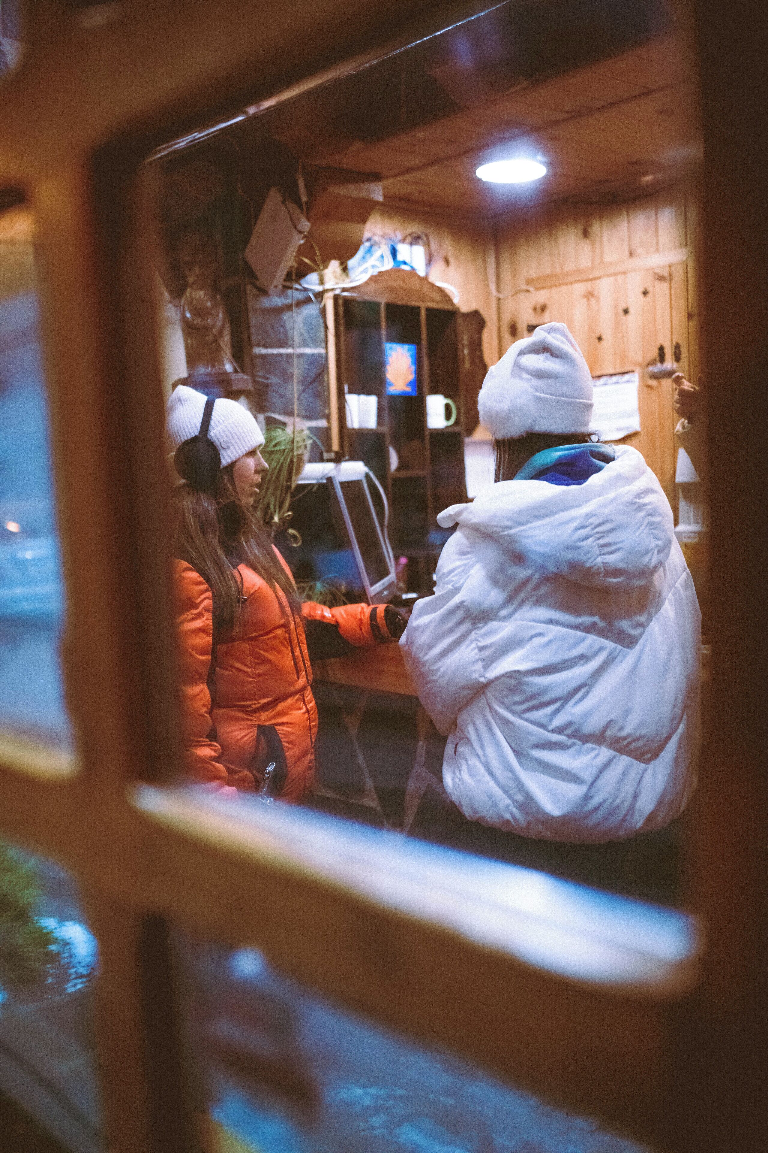 Un intérieur en bois où l’on voit une personne vêtue d’une blouse blanche et une autre personne vêtue d’un manteau orange, debout près d’un comptoir.