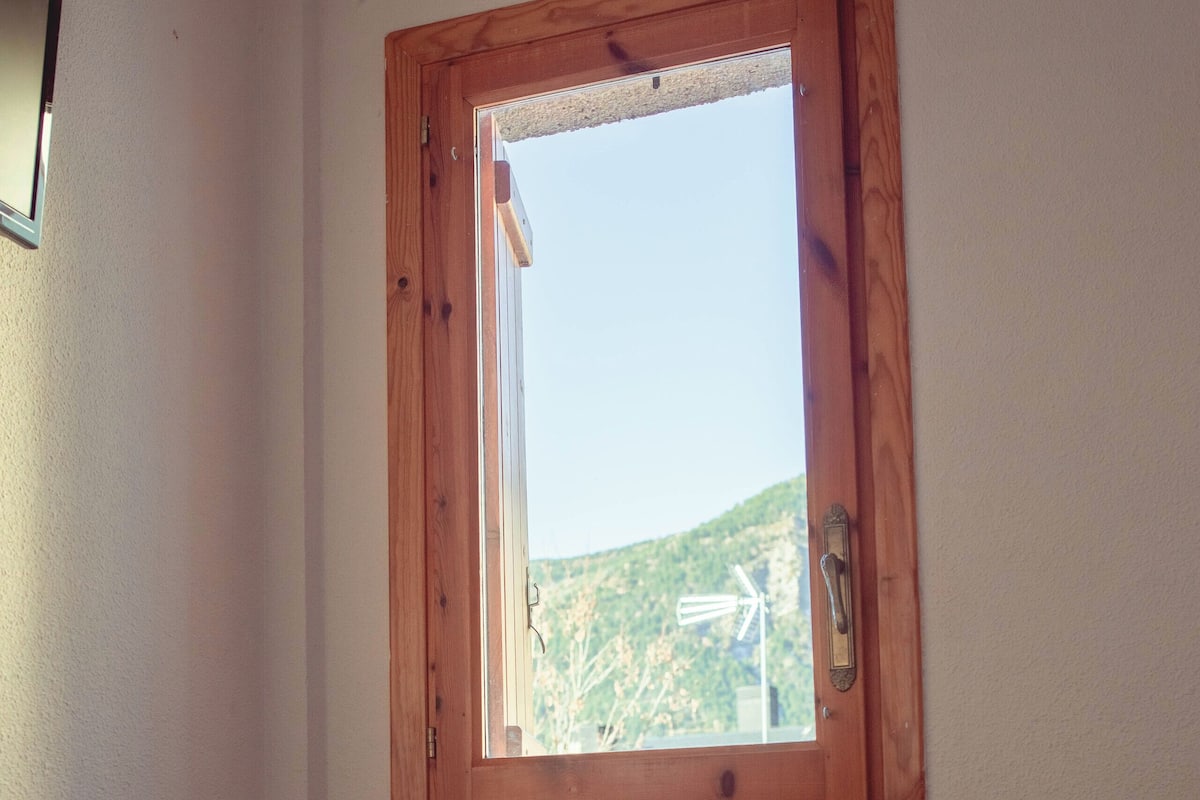 Ein Schlafzimmer mit einem hölzernen Kopfende, einem Einzelbett, einem Nachttisch mit einer kleinen Pflanze und einer Tasse, sowie einem Fenster mit Blick auf einen Berg und eine Windkraftanlage.