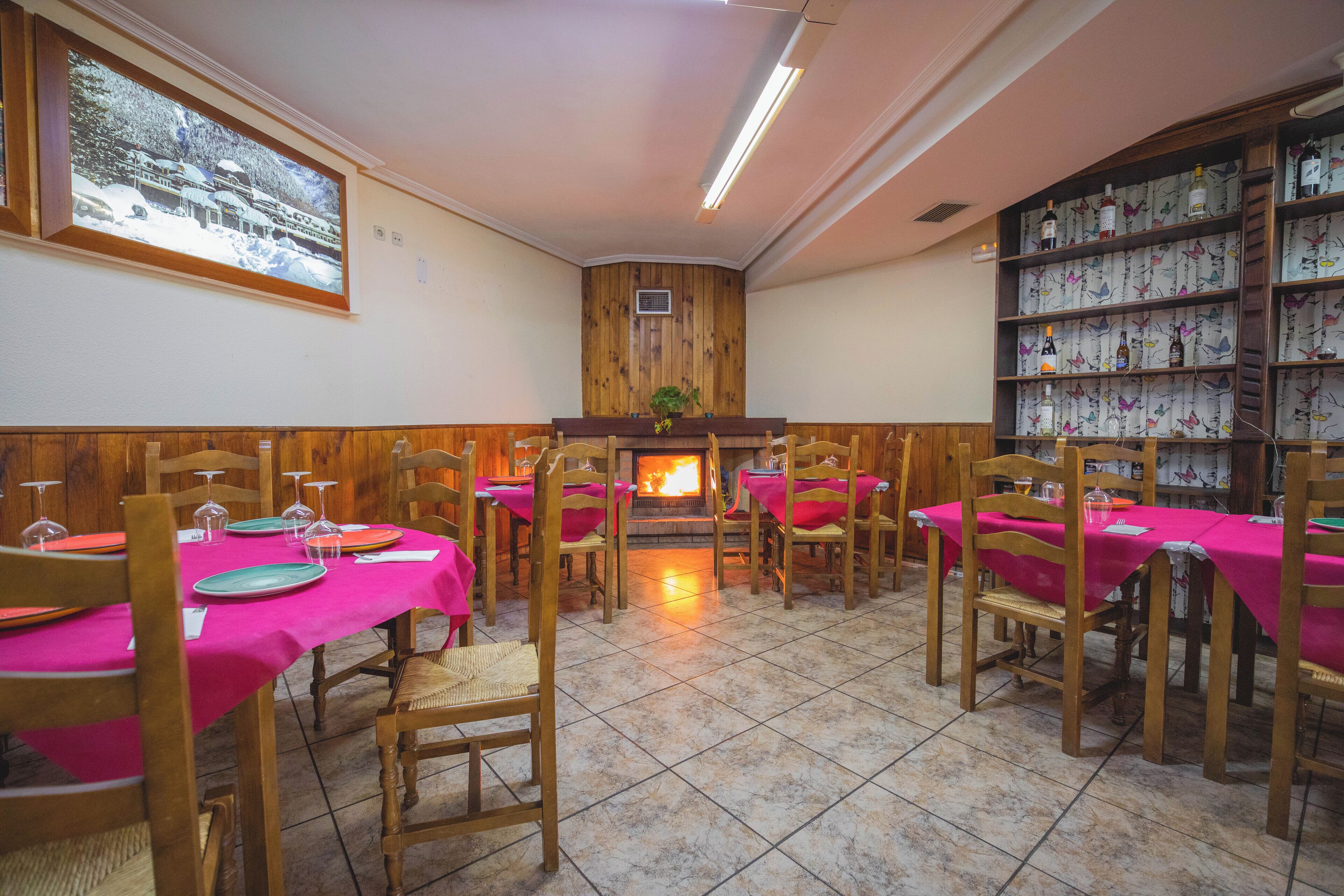 A dining area with wooden chairs and tables covered in pink tablecloths, a fireplace, and a wall-mounted display of bottles.