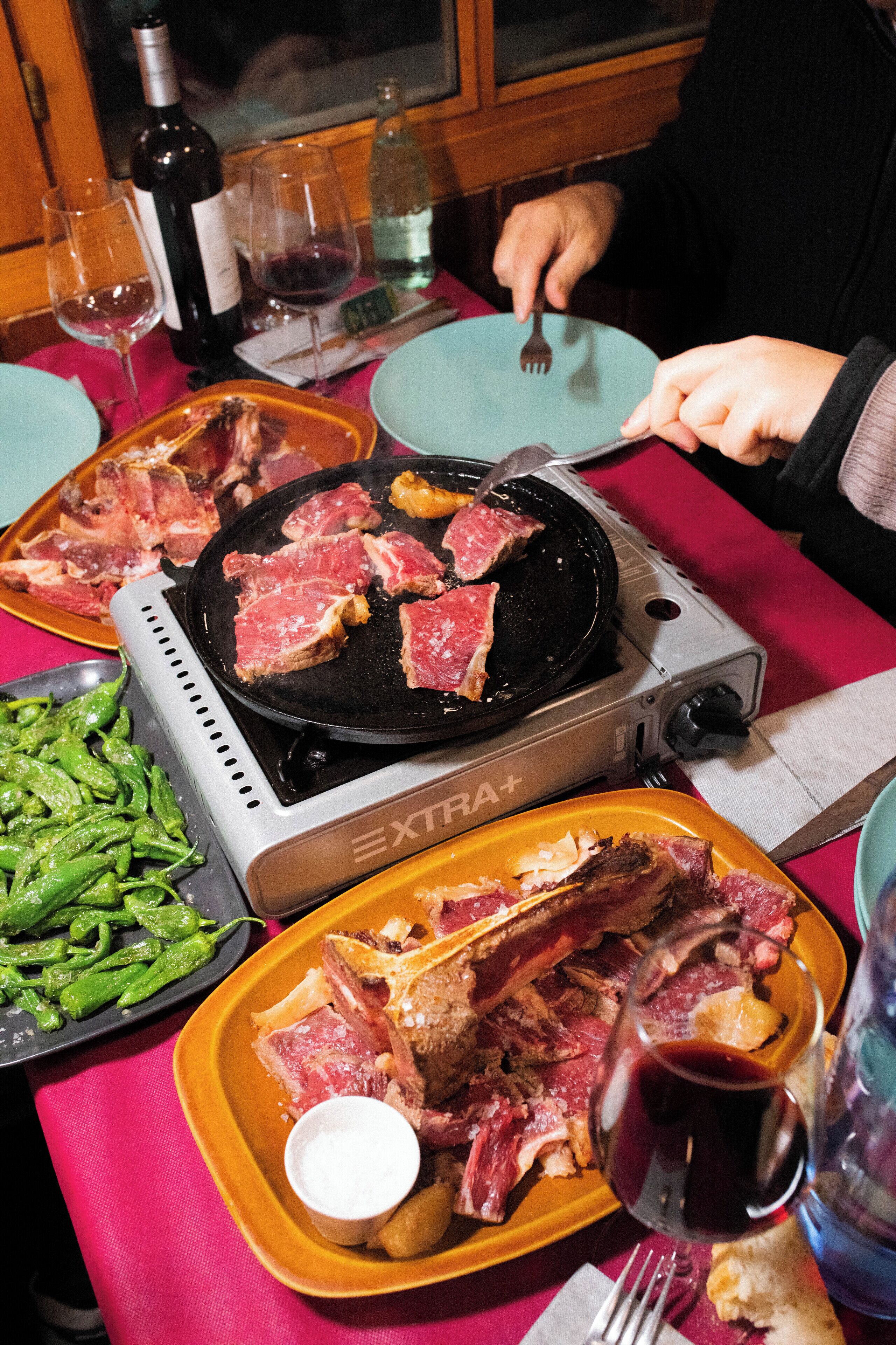 Una mesa de comedor con una parrilla portátil, platos de carne y copas de vino.