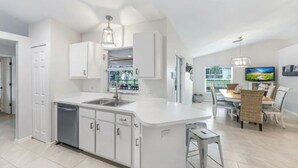 A modern kitchen with white cabinetry, a built-in dishwasher, and a dining area with wicker chairs and a TV. - Waterfront 3-Bed Home in SW Cape Heated Saltwater Pool, Boat Dock, Kayaks, Bikes (Cape Coral)