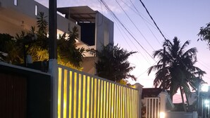 Ein modernes Haus mit einer hell erleuchteten, vertikal gestreiften Wand, einem verschlossenen Eingangsbereich und einer Palme im Hintergrund.