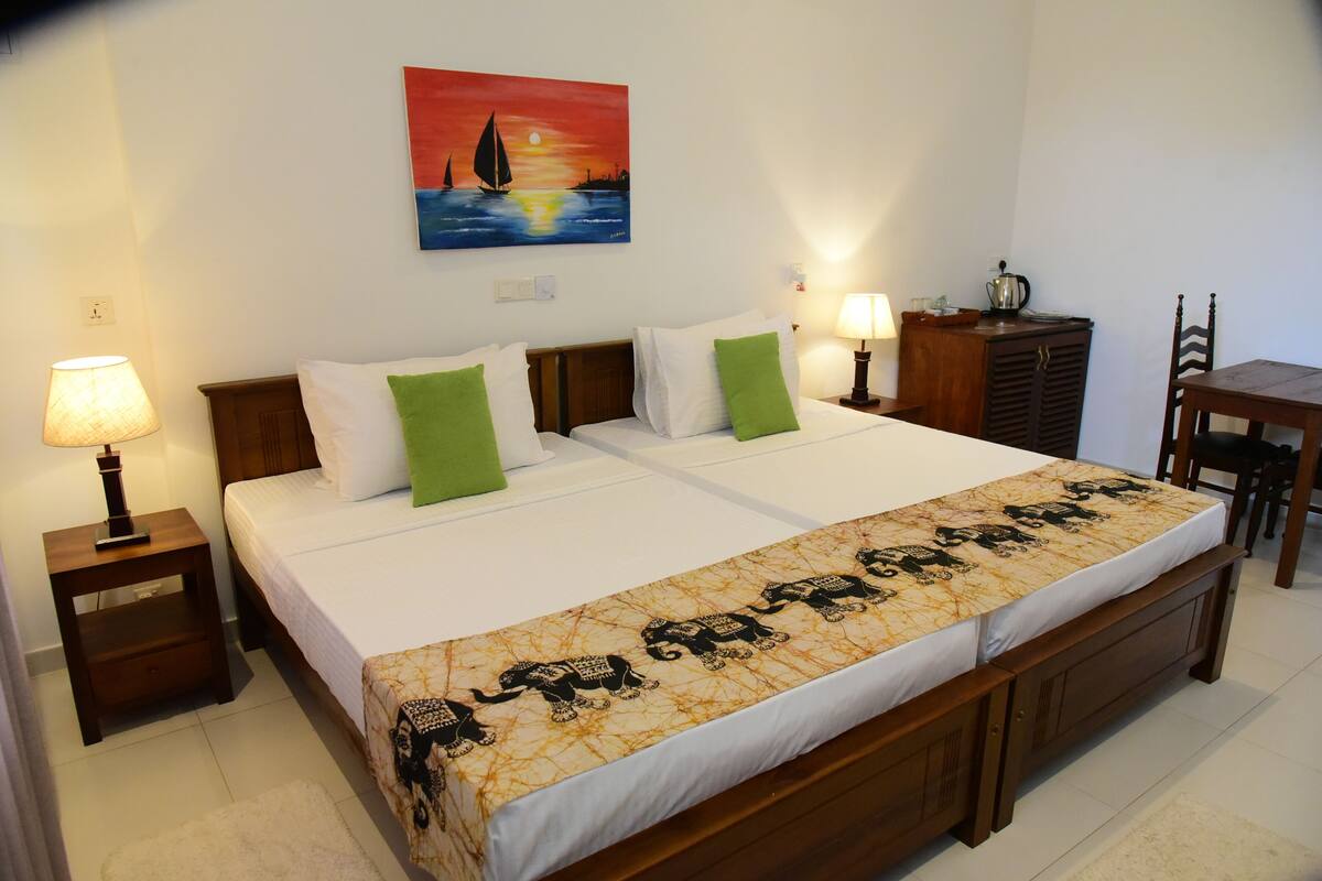 A double bed with a patterned bedspread, two bedside tables with lamps, a wooden nightstand, a wooden dresser, and a painting of a sailboat on the wall.