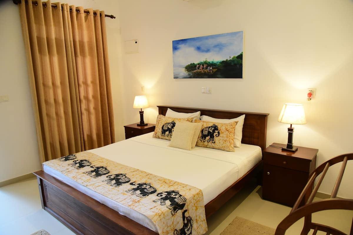 A neatly made bed with a patterned bedspread, two bedside lamps, a nightstand, a chair, and a painting on the wall.