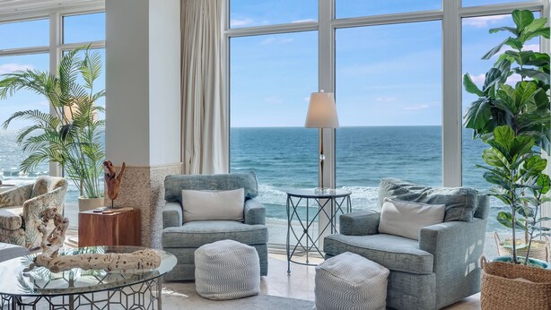 A spacious living room with a large window overlooking the ocean, featuring a grey sofa, ottomans, and a glass coffee table.