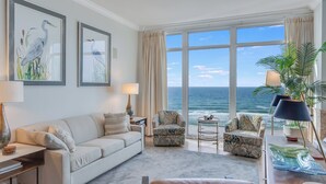A spacious living room with a large window overlooking the ocean, a sofa, two armchairs, and a coffee table.