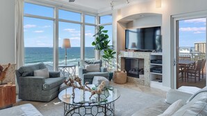 A spacious living room with large windows, a ceiling fan, a fireplace, and a television.