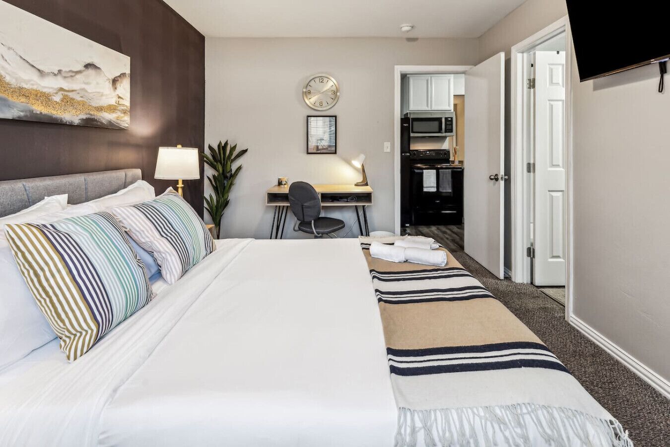 Hotellihuone, jossa on suuri sänky, työpöytä, tuoli, kello ja avoinna oleva ovi, joka johtaa keittiöön.