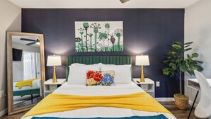 A bedroom with a bed, two bedside tables, a mirror, a green upholstered headboard, a wall painting, and a potted plant. - Prime 1BD Close to the Action (Arlington)