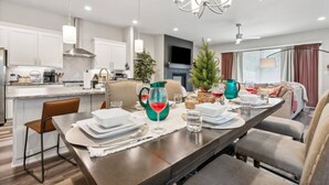 A modern kitchen with white cabinets, a dining area with a long table set for four, and a living area with a sofa and a TV. - Joyous 3BDTownhouse:  UltraCool & Spacious! (Fort Worth)