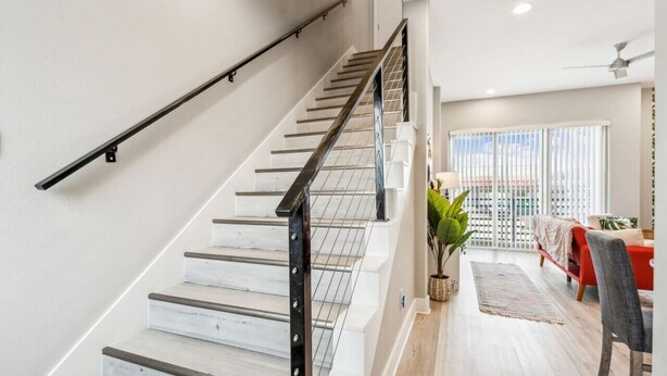 Un escalier avec une rampe métallique, des murs blancs et une vue sur un balcon agrémenté d’un canapé rouge et d’une plante.